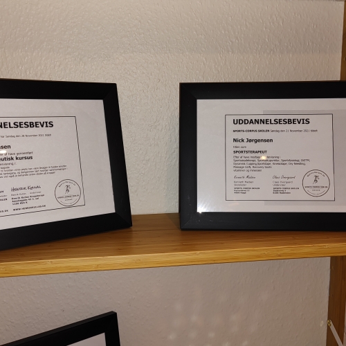 To indrammede diplomer fra massagekurser. Diplomerne viser certificering og ekspertise inden for massagebehandling, udstillet på en træhylde i et professionelt behandlerrum.