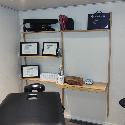 Et massagerum med en hylde fuld af udstyr, diplomer og plejeprodukter. Rummet er indrettet med en sort massagebriks og en minimalistisk stil. Perfekt til afslappende massagebehandlinger
