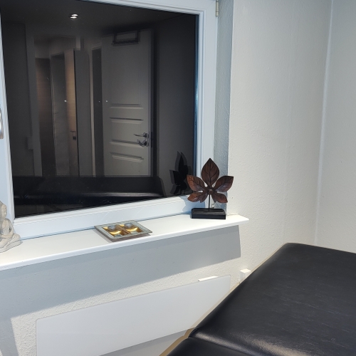 Et vindue i et massagerum med udsigt til gangen. Vindueskarmen er dekoreret med små ornamenter, herunder en Buddha-figur og en træskulptur, der skaber en afslappende atmosfære.