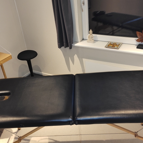 En sort massagebriks placeret i et hyggeligt behandlerrum. I baggrunden ses en træhylde med plejeprodukter og et vindue dekoreret med en Buddha-figur og en bladformet skulptur, som skaber en afslappende atmosfære til massagebehandlinger.