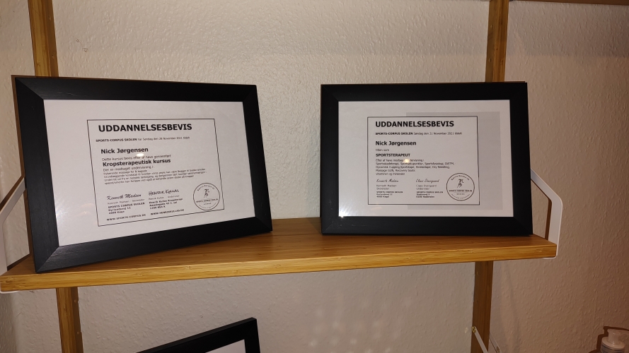 To indrammede diplomer fra massagekurser. Diplomerne viser certificering og ekspertise inden for massagebehandling, udstillet på en træhylde i et professionelt behandlerrum.