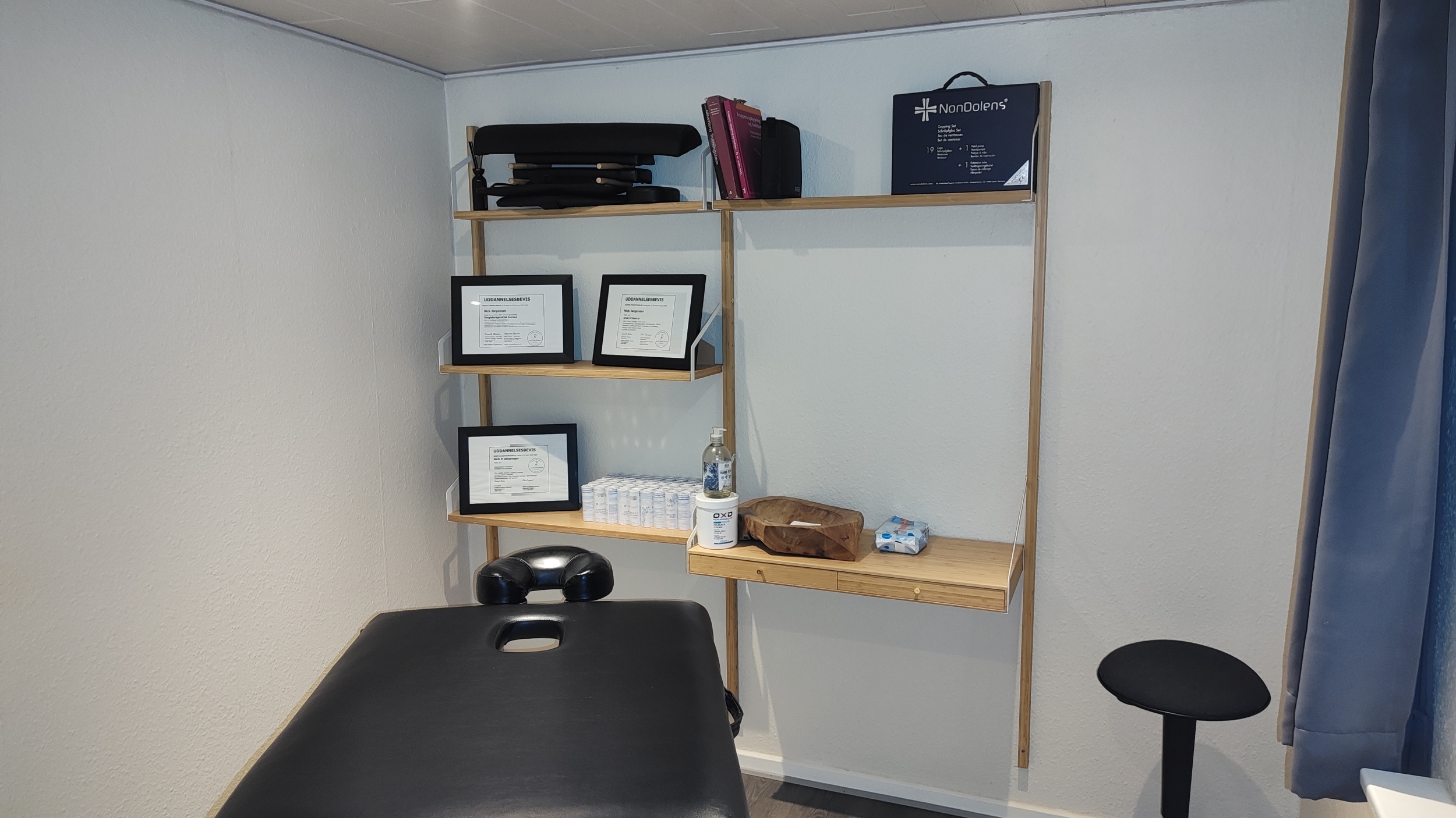 Et massagerum med en hylde fuld af udstyr, diplomer og plejeprodukter. Rummet er indrettet med en sort massagebriks og en minimalistisk stil. Perfekt til afslappende massagebehandlinger