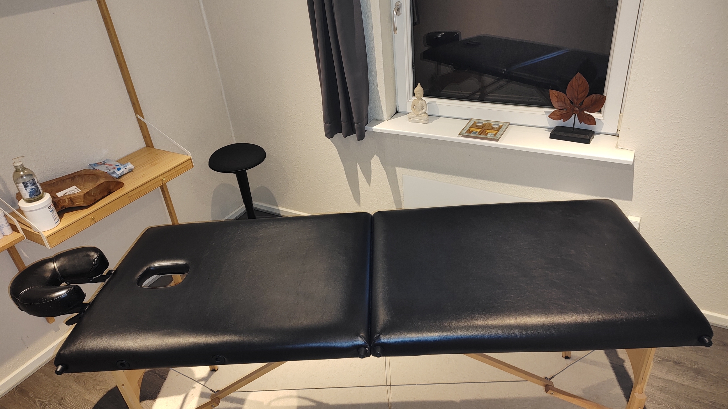 En sort massagebriks placeret i et hyggeligt behandlerrum. I baggrunden ses en træhylde med plejeprodukter og et vindue dekoreret med en Buddha-figur og en bladformet skulptur, som skaber en afslappende atmosfære til massagebehandlinger.
