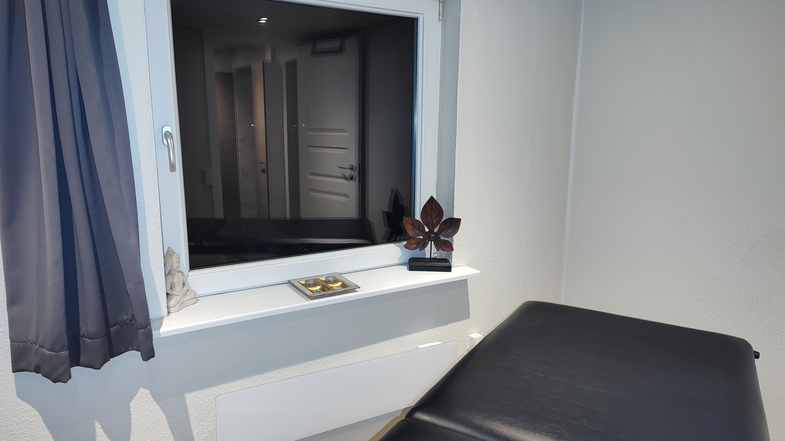 Et vindue i et massagerum med udsigt til gangen. Vindueskarmen er dekoreret med små ornamenter, herunder en Buddha-figur og en træskulptur, der skaber en afslappende atmosfære.